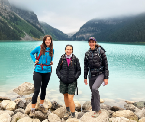 Photo of three people standing in front of a crystal clear lake with mountains in the background on a hike