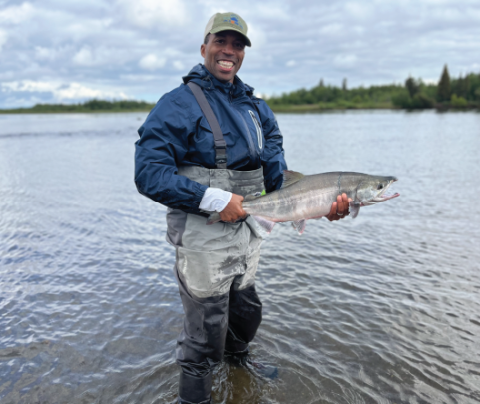 Photo of Baratunde Cola wearing waders, standing in a body of water holding a large fish, and smiling.