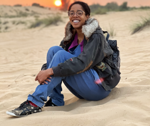 Photo of Madison Maxey sitting in sand at the beach in a jacket and jeans with the sun setting behind her