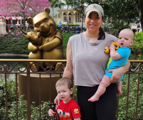 Photo of Carley Mollica holding her infant son standing next to her toddler aged son with a bronze Winnie the Pooh and Piglet statue in the background.