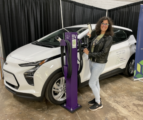 Photo of Helena Cristobal standing in front of an electric vehicle charging station with an electric vehicle behind her